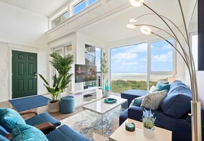 A modern living room with a large window, a flat-screen TV, and a blue sofa. - Amazing Oceanfront Views! Steps to the Sand! Rockaway 2BR Condo Nedonna Beach! (Rockaway Beach)