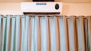 A white air conditioning unit with a digital display and speed control buttons above a striped curtain. - Hotel Talon (Mumbai)