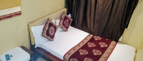 A neatly made bed with a patterned bedspread and two decorative pillows, a bedside table with a folded white cloth and a small electronic device, and a dark curtain covering a window.