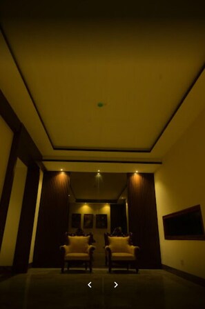 Un pasillo tenuemente iluminado con dos sillones enfrentados, una lámpara en el techo y un cuadro en la pared.