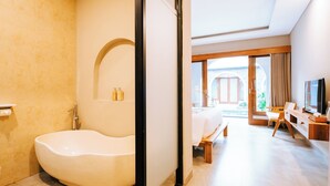 A modern bathroom with a freestanding bathtub, a vanity with a mirror, and a shower area. The room has a large window providing natural light and a view of the outdoors. - villa koura kuta (Kuta)