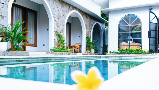 A modern poolside area with a stone facade, arched doorways, and a clear pool reflecting the surroundings. - villa koura kuta (Kuta)