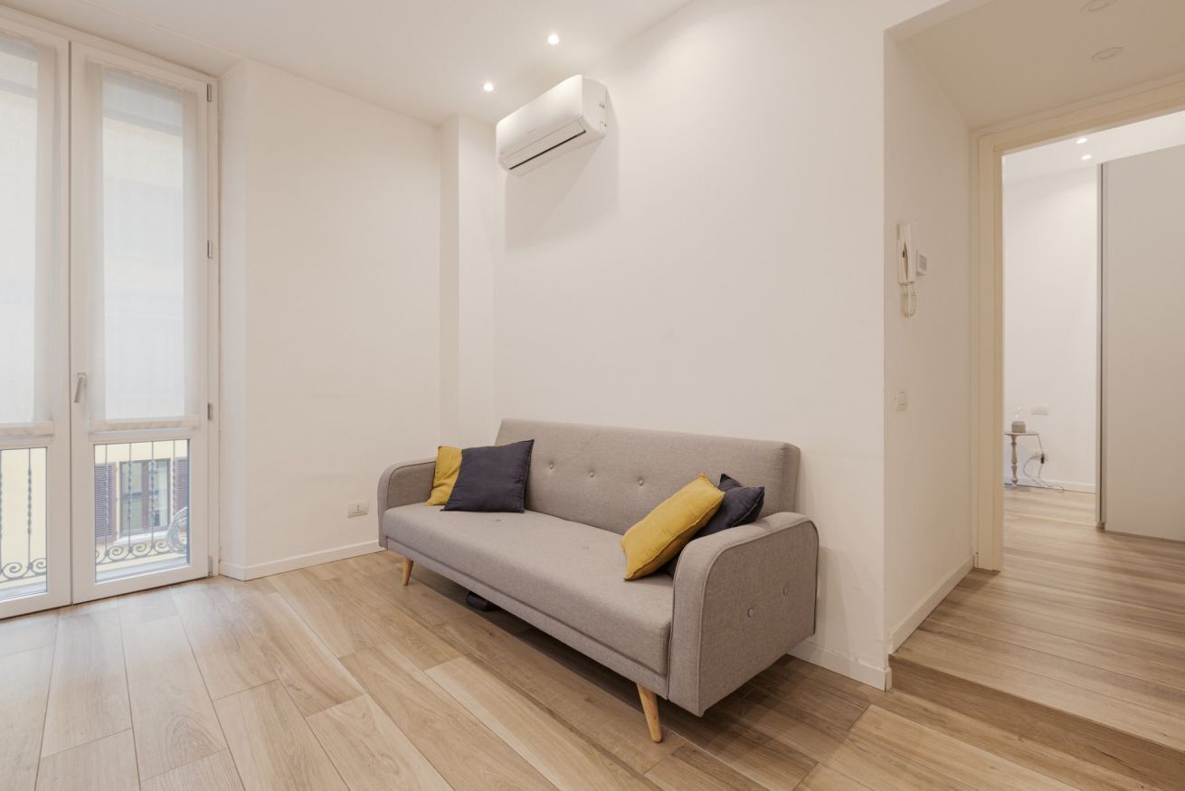 A modern living room with a grey sofa, a white air conditioning unit, and a wooden floor.