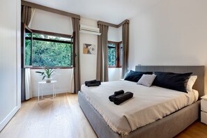 Un dormitorio con una cama grande, una mesita con una planta y una ventana con cortinas.