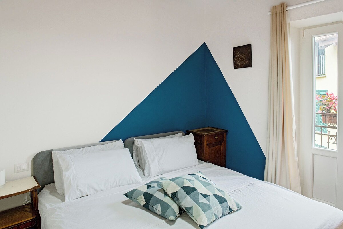A bedroom with a bed, white pillows, a geometric-patterned pillow, a nightstand, a small wall decoration, and a window with curtains.