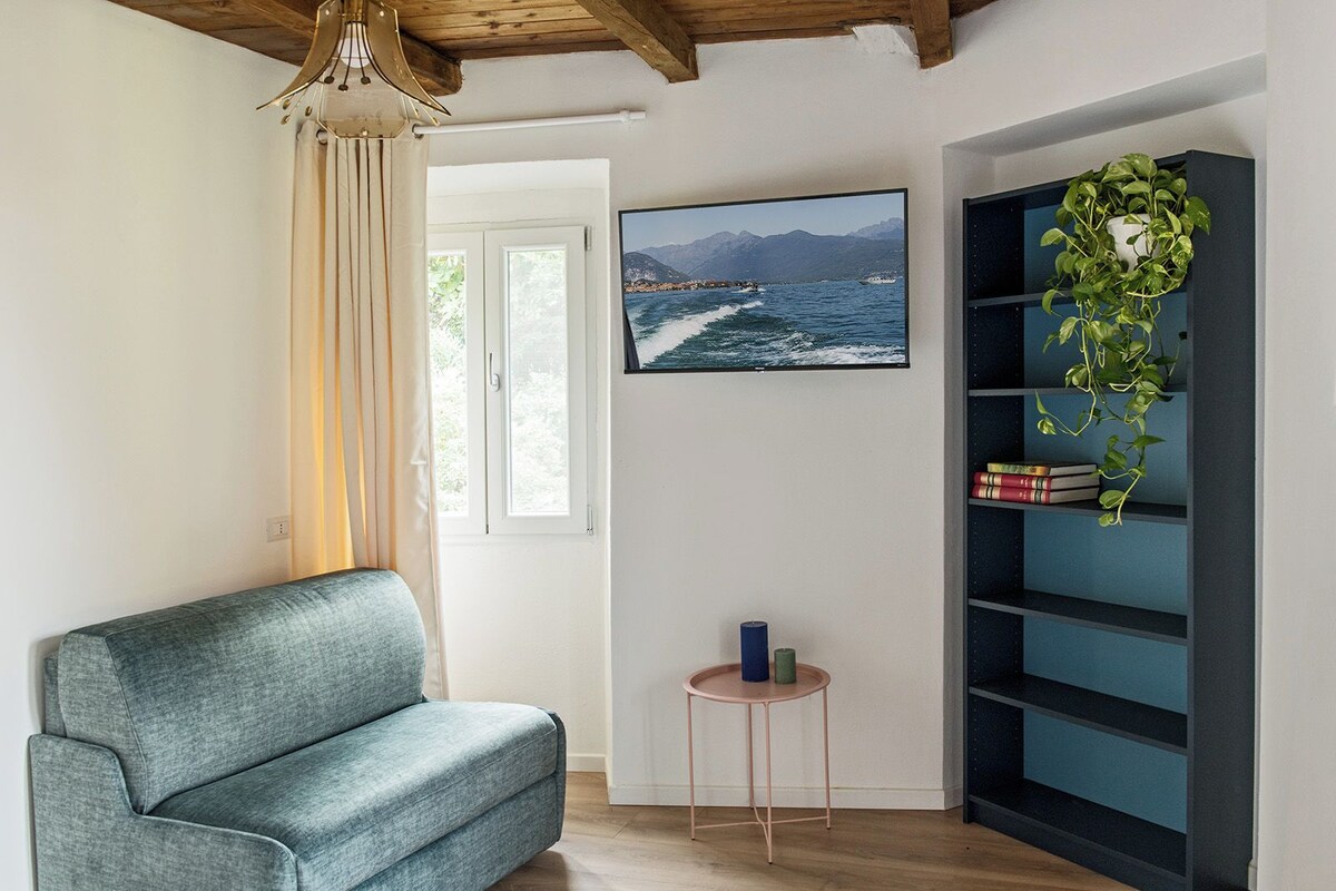 Uma sala de estar com um sofá, uma mesa redonda pequena, uma estante de livros e uma televisão grande exibindo uma paisagem costeira.