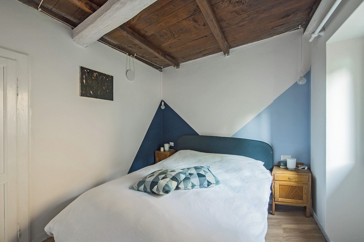 A bedroom with a sloped ceiling, a bed with a blue headboard, a bedside table with a lamp, and a wall-mounted light fixture.