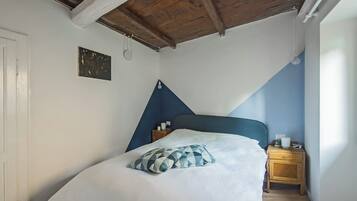 Un dormitorio con techo inclinado, una cama con cabecera azul, una mesita de noche con lámpara y un aplique de pared.