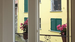 Un balcón con barandilla metálica, un edificio amarillo con contraventanas verdes y macetas con flores rosadas.
