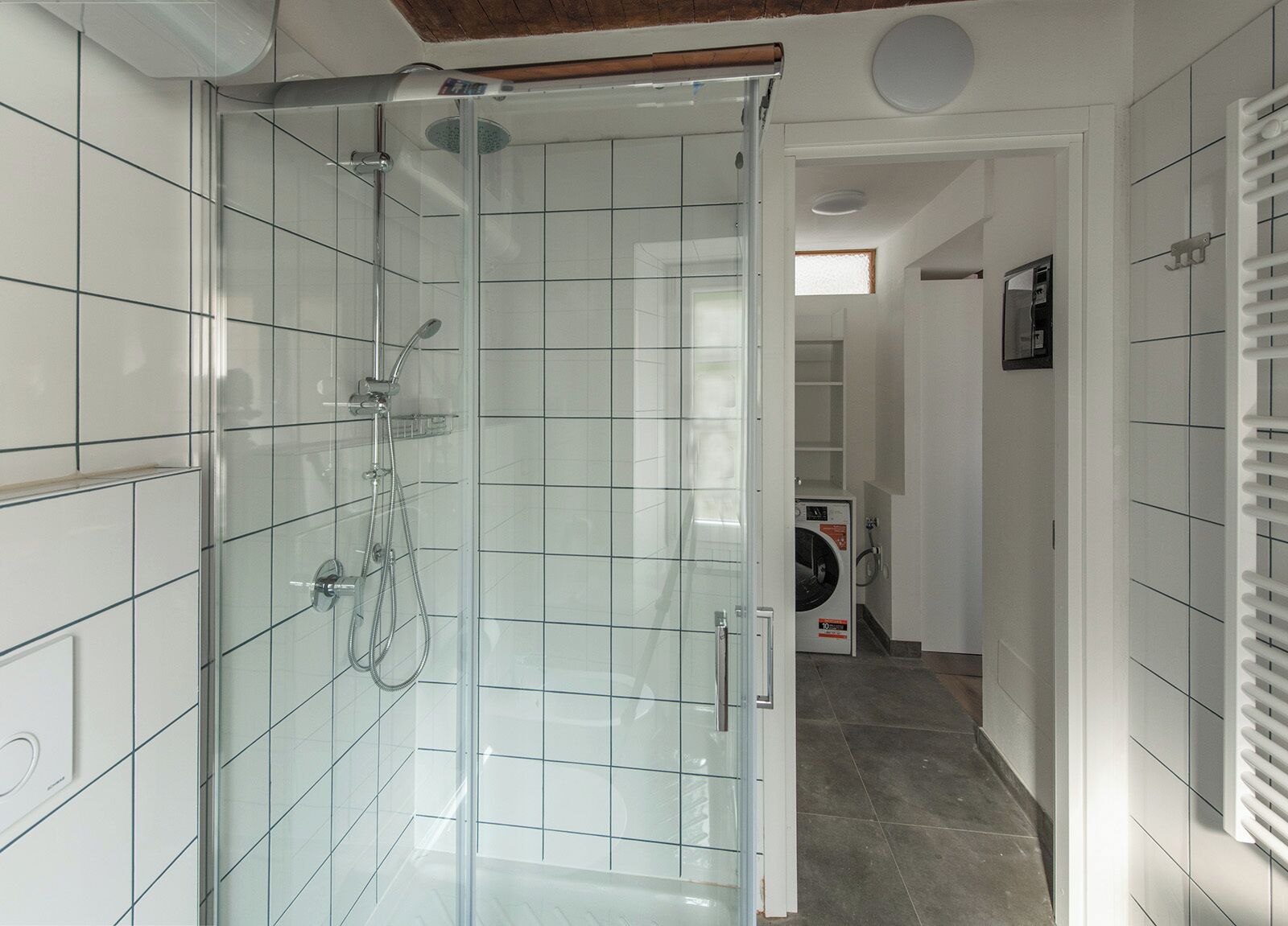 A modern bathroom with a glass-enclosed shower, white tiled walls, and a visible laundry area with a washing machine.