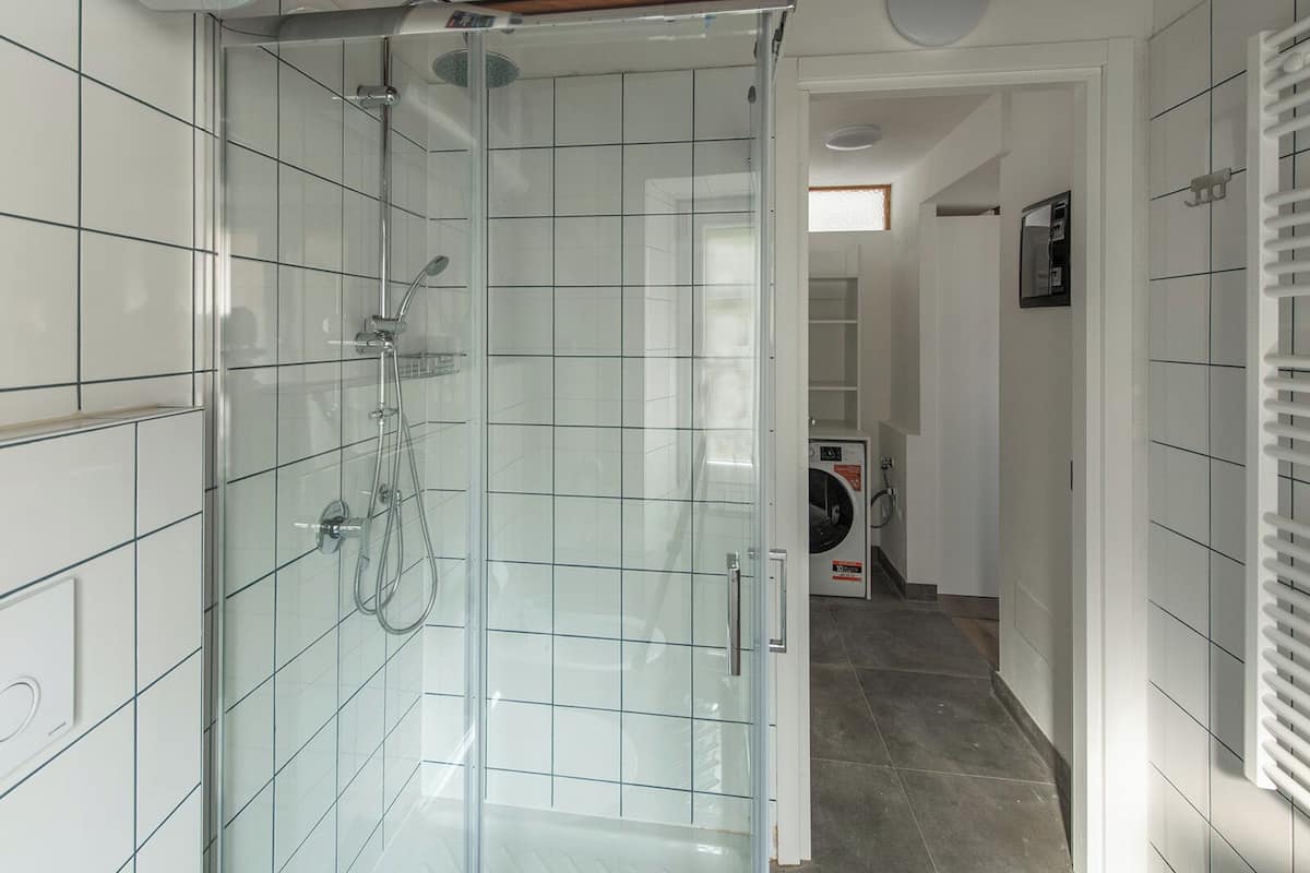 A modern bathroom with a glass-enclosed shower, white tiled walls, and a visible laundry area with a washing machine.