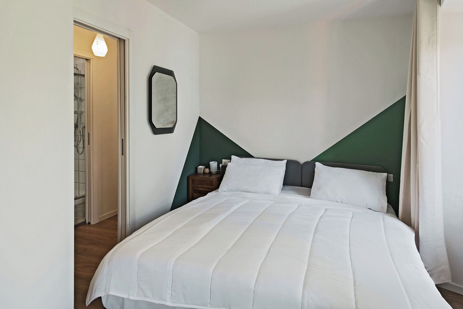 Una cama bien hecha con sábanas blancas, una mesita de noche con lámpara y una pared de acento verde.