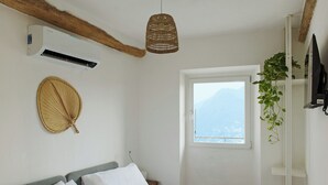 Un dormitorio con cama, aire acondicionado, un tapiz de mimbre, ventana con vistas a montañas y una planta sobre una estantería.