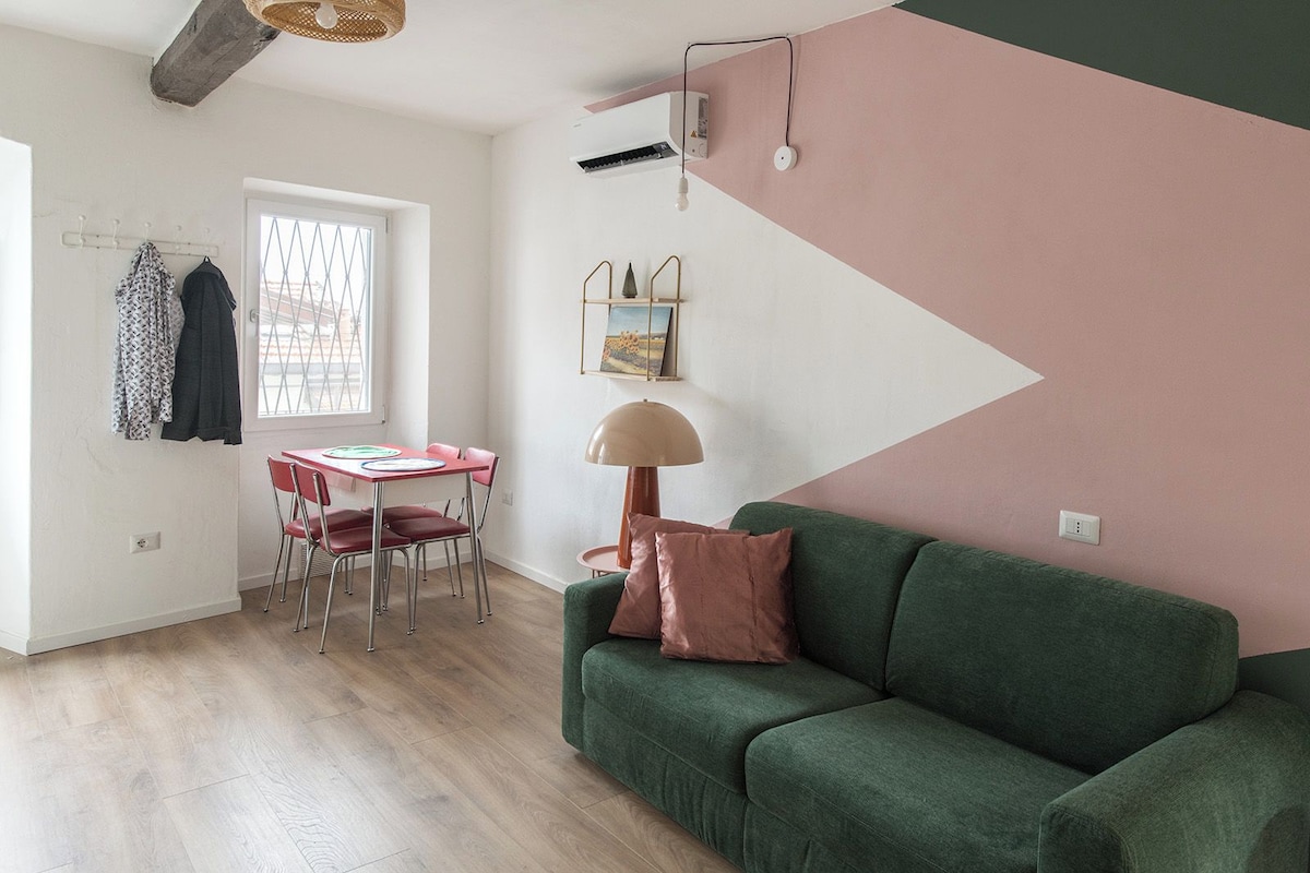 A room with a green sofa, a dining table with chairs, a window with a view, and a wall with a geometric design.