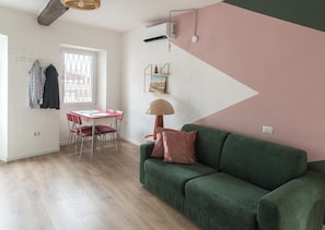 Quarto com sofá verde, mesa de jantar com cadeiras, janela com vista e parede com design geométrico.