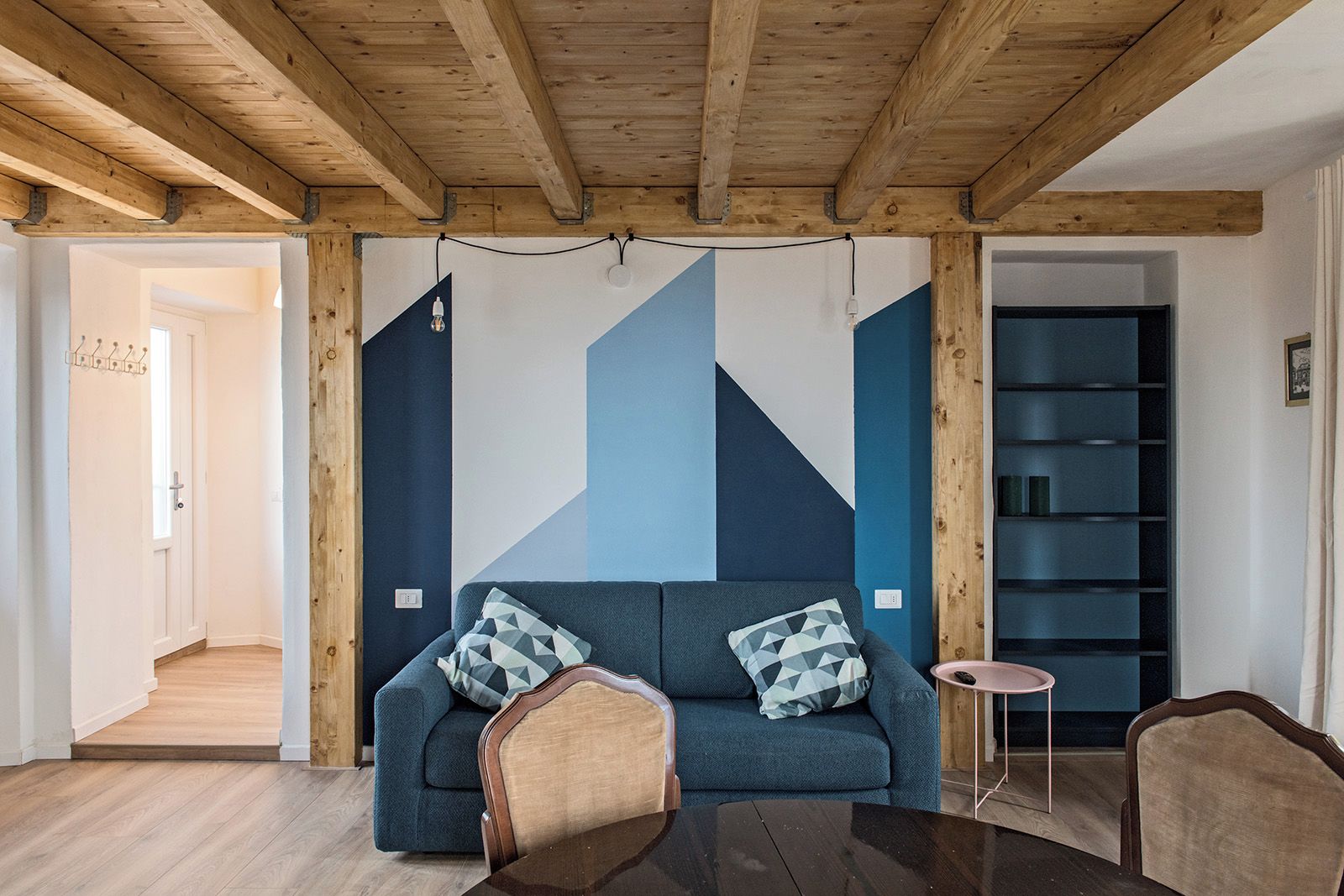 A modern living room with a blue sofa, wooden beams, a small round table, and a bookshelf.