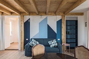 A modern living room with a blue sofa, wooden beams, a small round table, and a bookshelf. - Stresa Complex Levoasi (Stresa)
