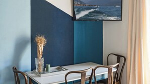 Un comedor con una mesa, sillas, un jarrón con plantas secas, una vela y un televisor en la pared mostrando una escena costera.