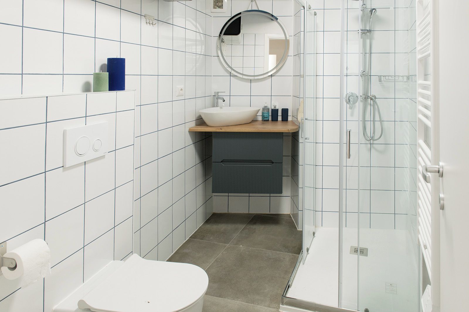 Un baño moderno con paredes de azulejos blancos, un espejo redondo, un lavabo con encimera de madera, un inodoro y una zona de ducha con mampara de vidrio.