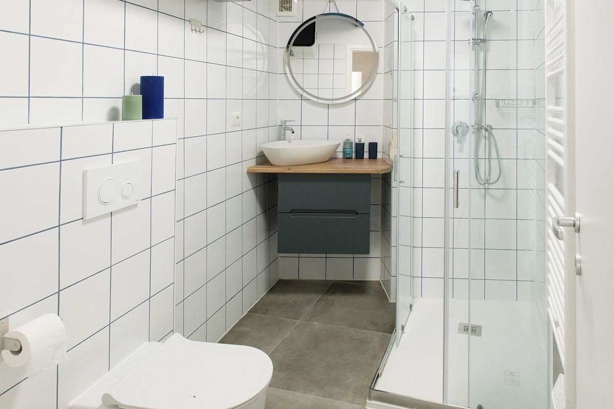 A modern bathroom with white tiled walls, a round mirror, a sink with a wooden countertop, a toilet, and a glass-enclosed shower area.