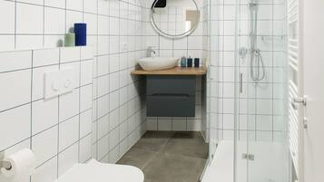 Un baño moderno con paredes de azulejos blancos, un espejo redondo, un lavabo con encimera de madera, un inodoro y una zona de ducha con mampara de vidrio.