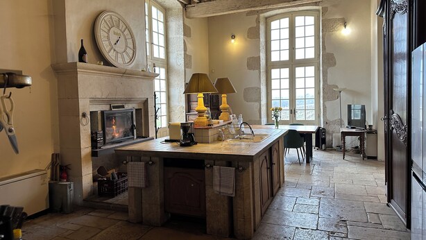 A rustic kitchen with a fireplace, a large clock, and a stone floor.