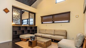 A living room with a sofa, a coffee table, a bench, a decorative wall piece, and a window with blinds. - 1688 House Churaumi (Motobu)