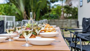 A wooden table set with white plates, glasses of white wine, and a bottle of wine, surrounded by chairs and greenery in the background. - 1688 House Churaumi (Motobu)