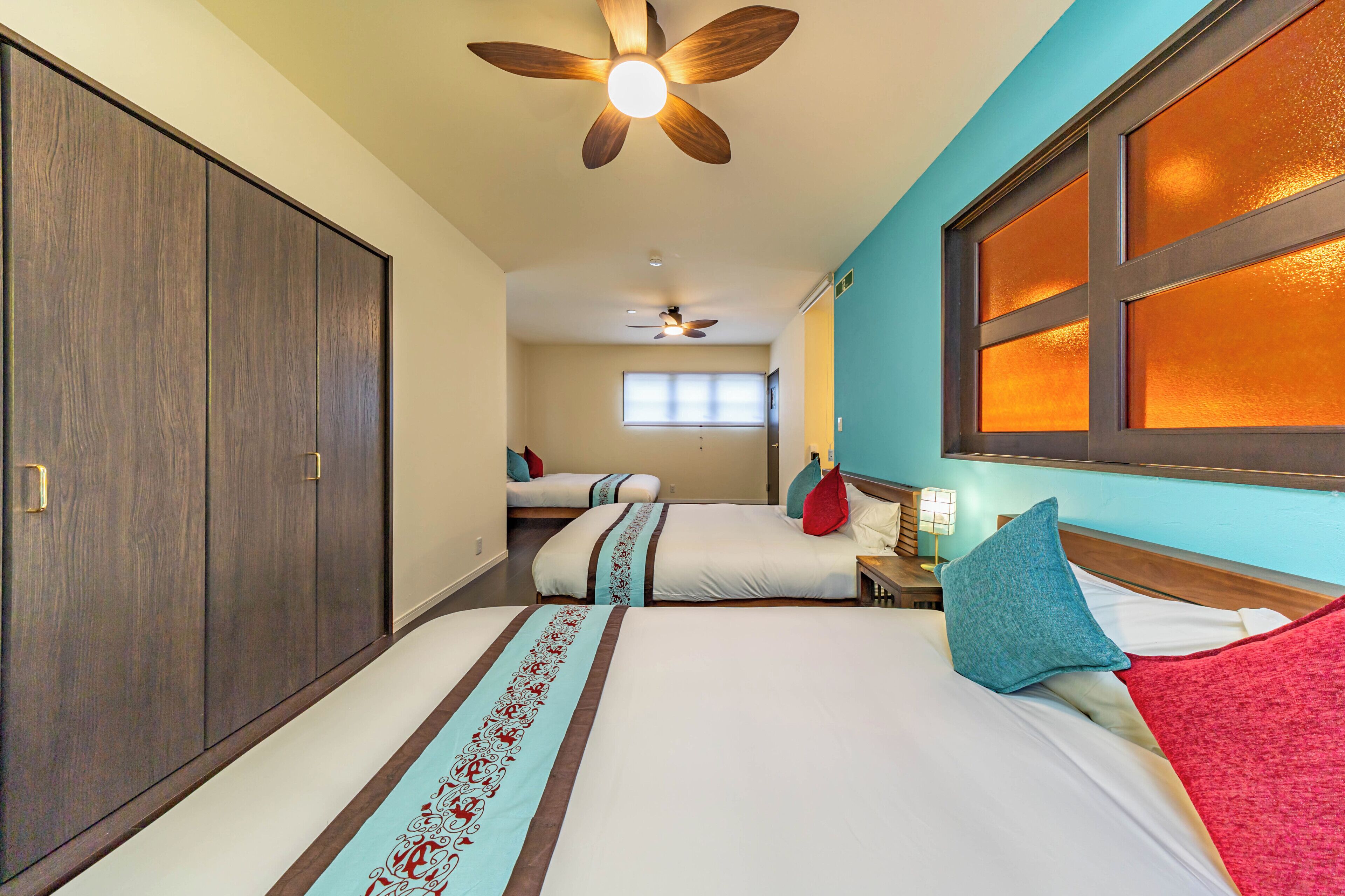 A hotel room with two beds, a ceiling fan, a window with blinds, and a nightstand with a lamp.