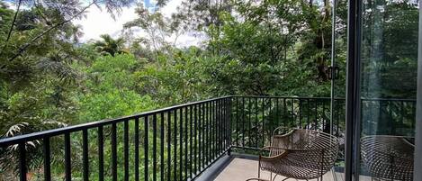A balcony with a metal railing, two wicker chairs, and a view of dense greenery and trees.