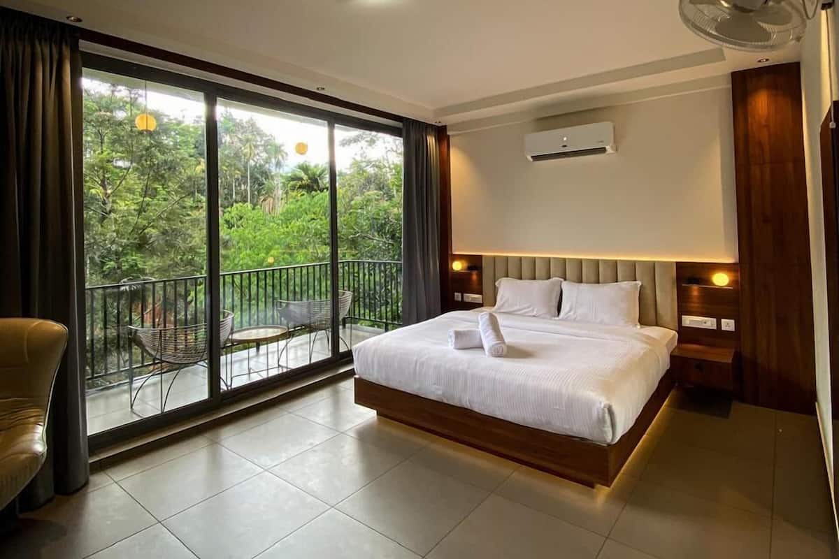 Quarto de hotel moderno com uma cama grande, uma varanda com cadeira e vista para áreas verdes.