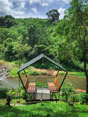 A hexagonal wooden gazebo with benches, overlooking a river and lush greenery. - Rill woods (Vythiri)