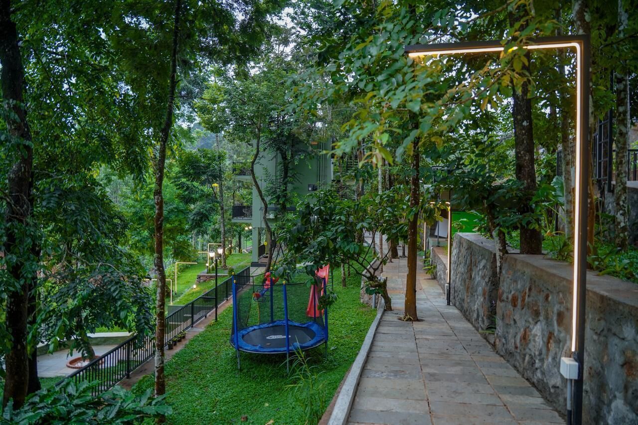 A playground with a trampoline and slides, surrounded by trees and a stone wall.