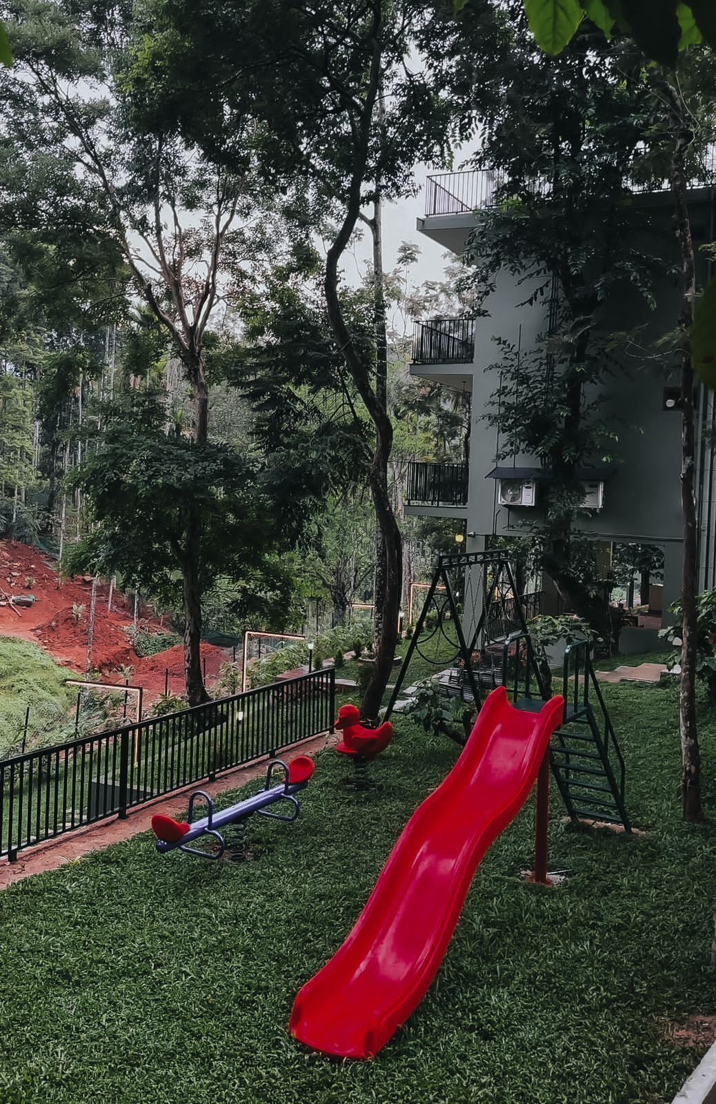 Un parque infantil con un tobogán rojo y columpios, rodeado de árboles y un edificio al fondo.