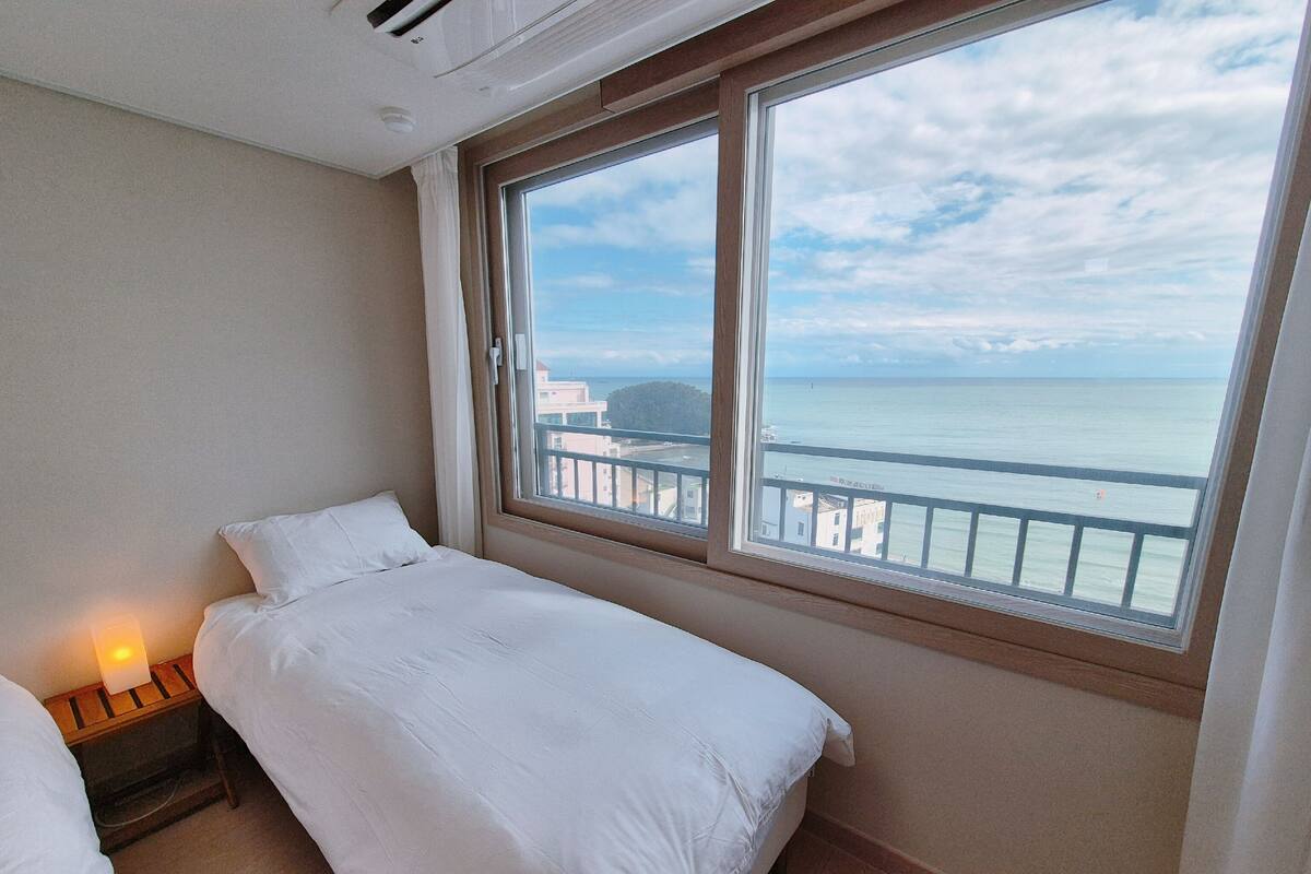 A hotel room with a bed, a nightstand with a lamp, and a large window offering a view of the sea and buildings.