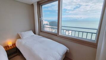 A hotel room with a bed, a nightstand with a lamp, and a large window offering a view of the sea and buildings.