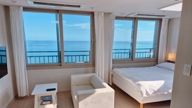 A hotel room with a bed, a small table, a chair, and large windows offering a view of the sea. - The First Ocean Songjung (Busan)