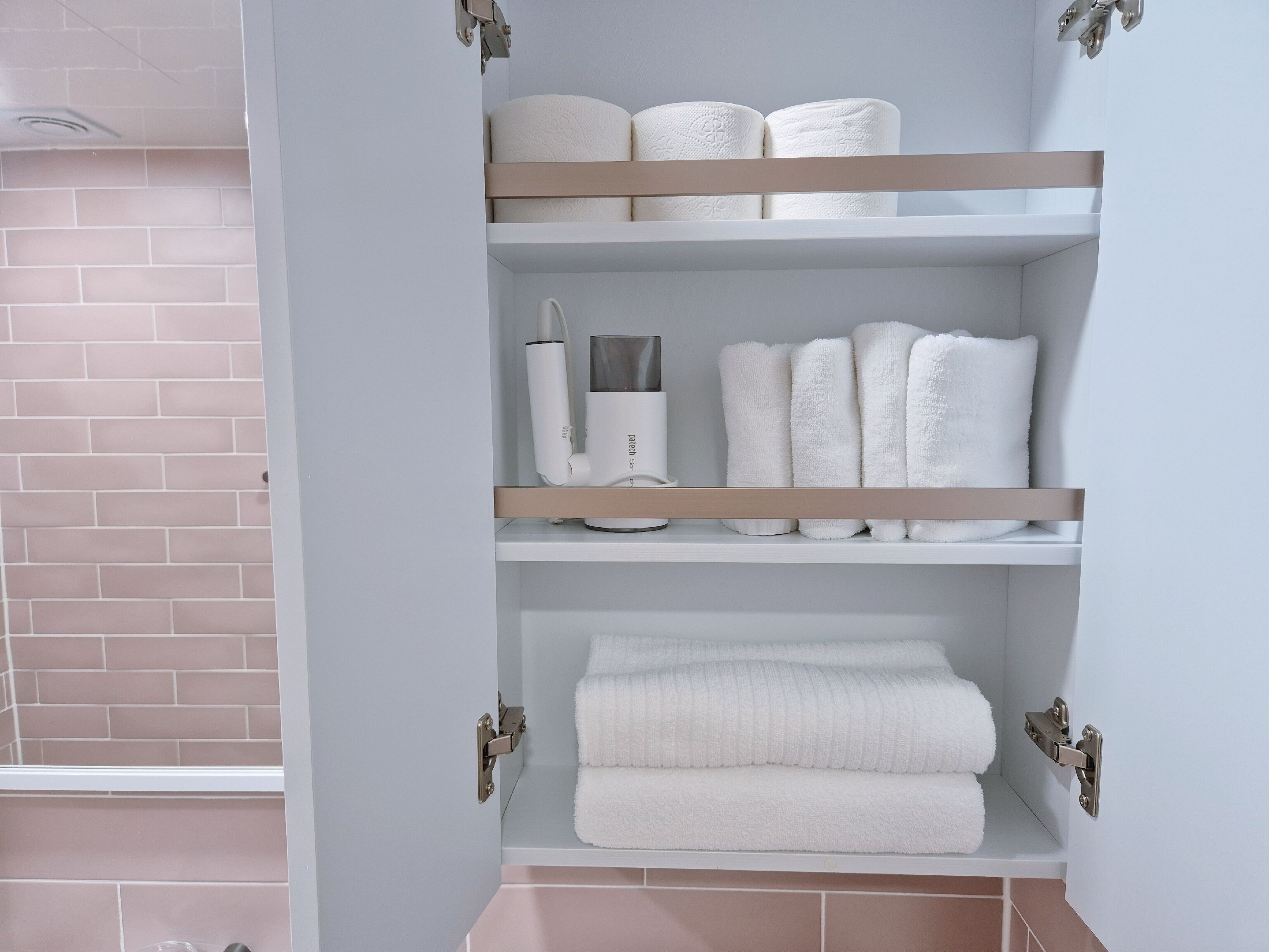 Un baño con pared de azulejos blancos, pared de ladrillo y estante con toallas y un secador de pelo.