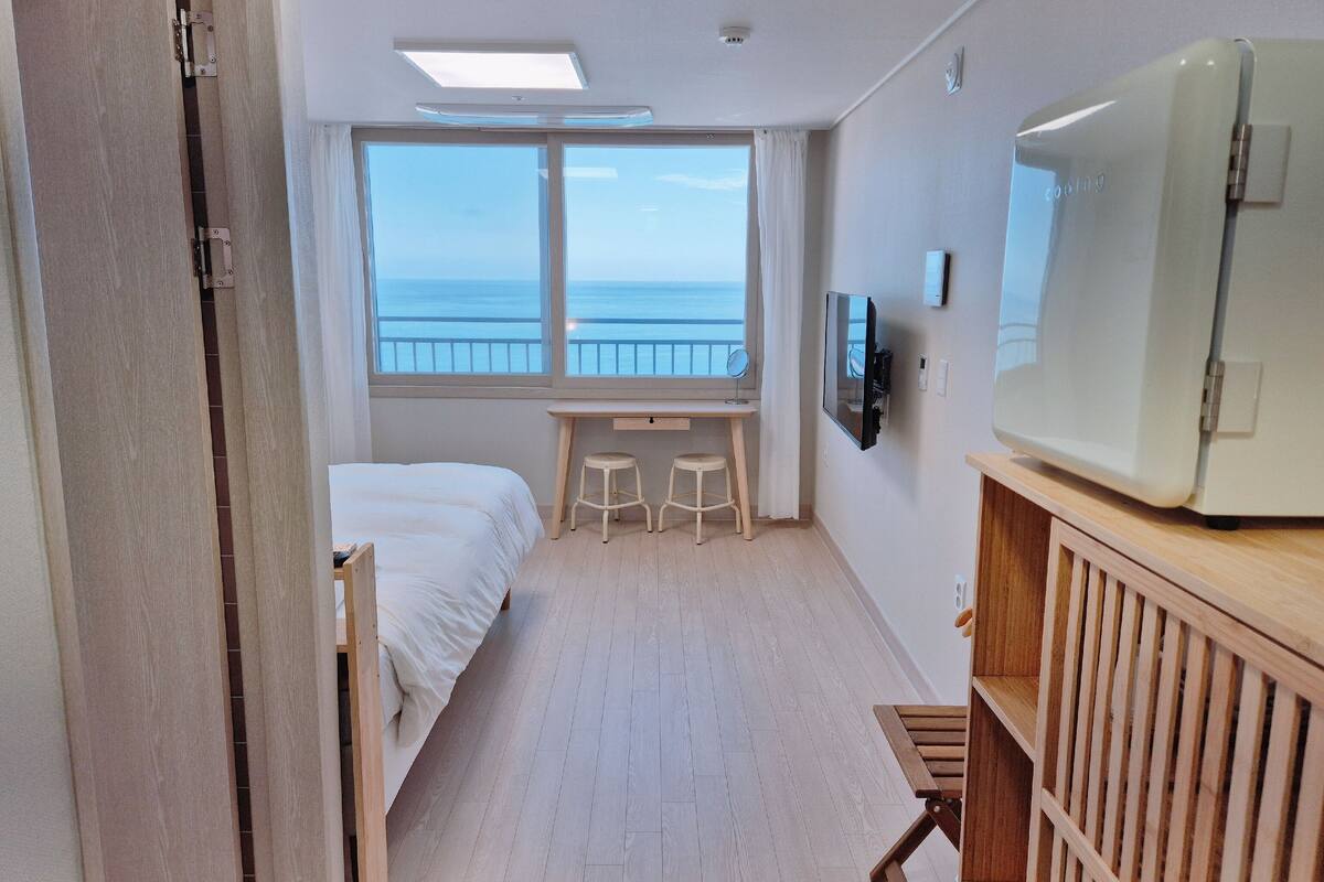 A hotel room with a bed, a desk, two stools, a TV, and a view of the sea.