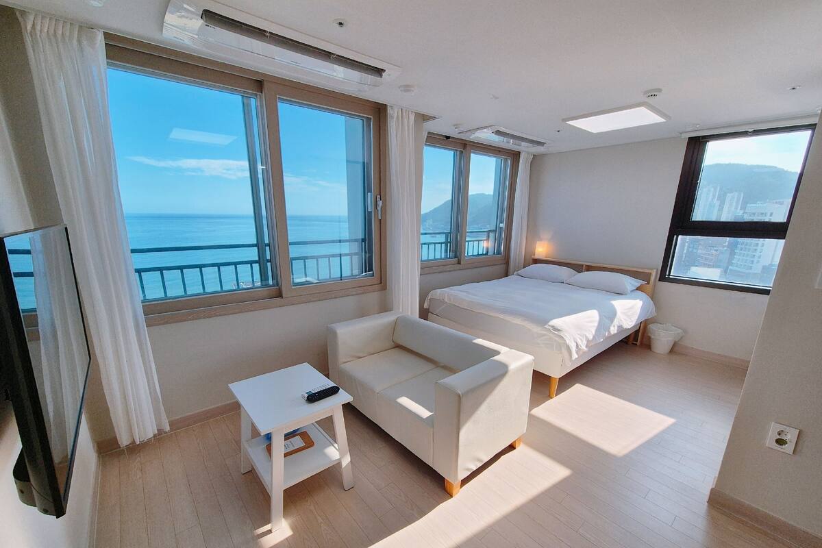 A modern hotel room with a bed, a sofa, a small table, and large windows offering a view of the sea.