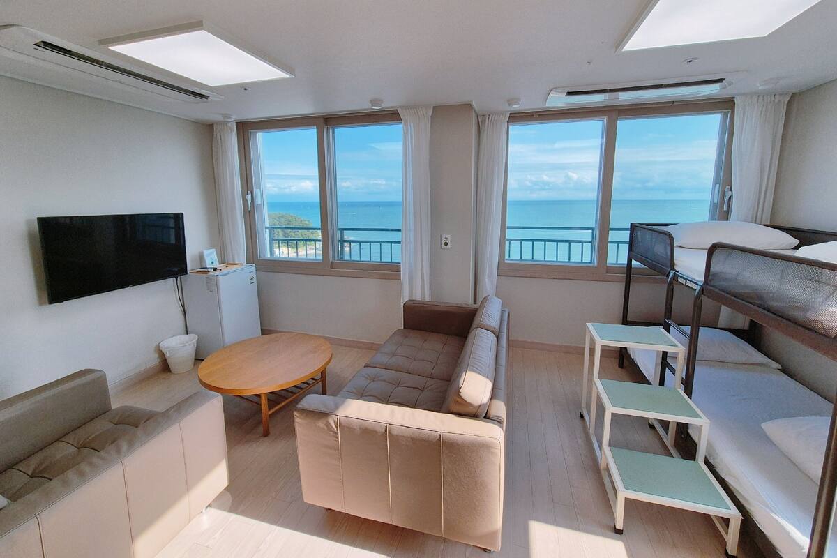 A compact room with a sofa, a small table, a bunk bed, a TV, and large windows offering a view of the sea.