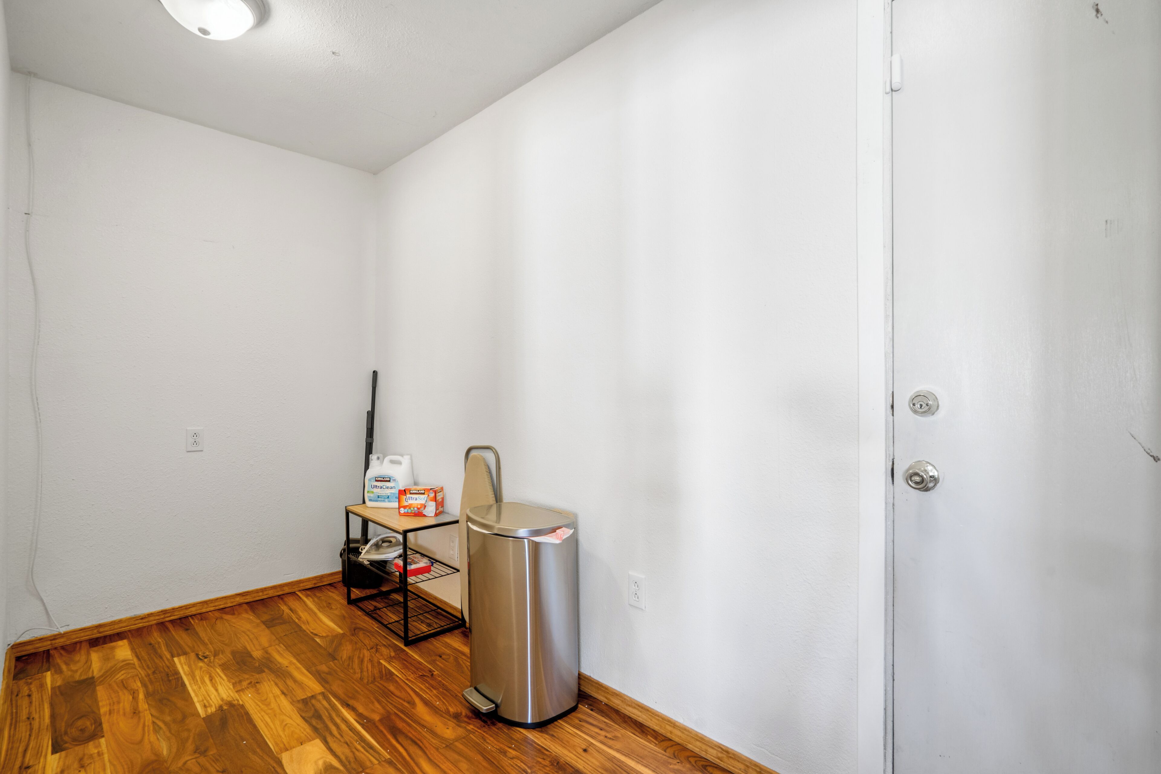 A room with white walls, a wooden floor, a trash can, a small table with a chair, and a door.