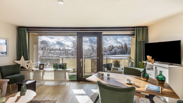 A modern living room with a large window, a flat-screen TV, and a dining area.