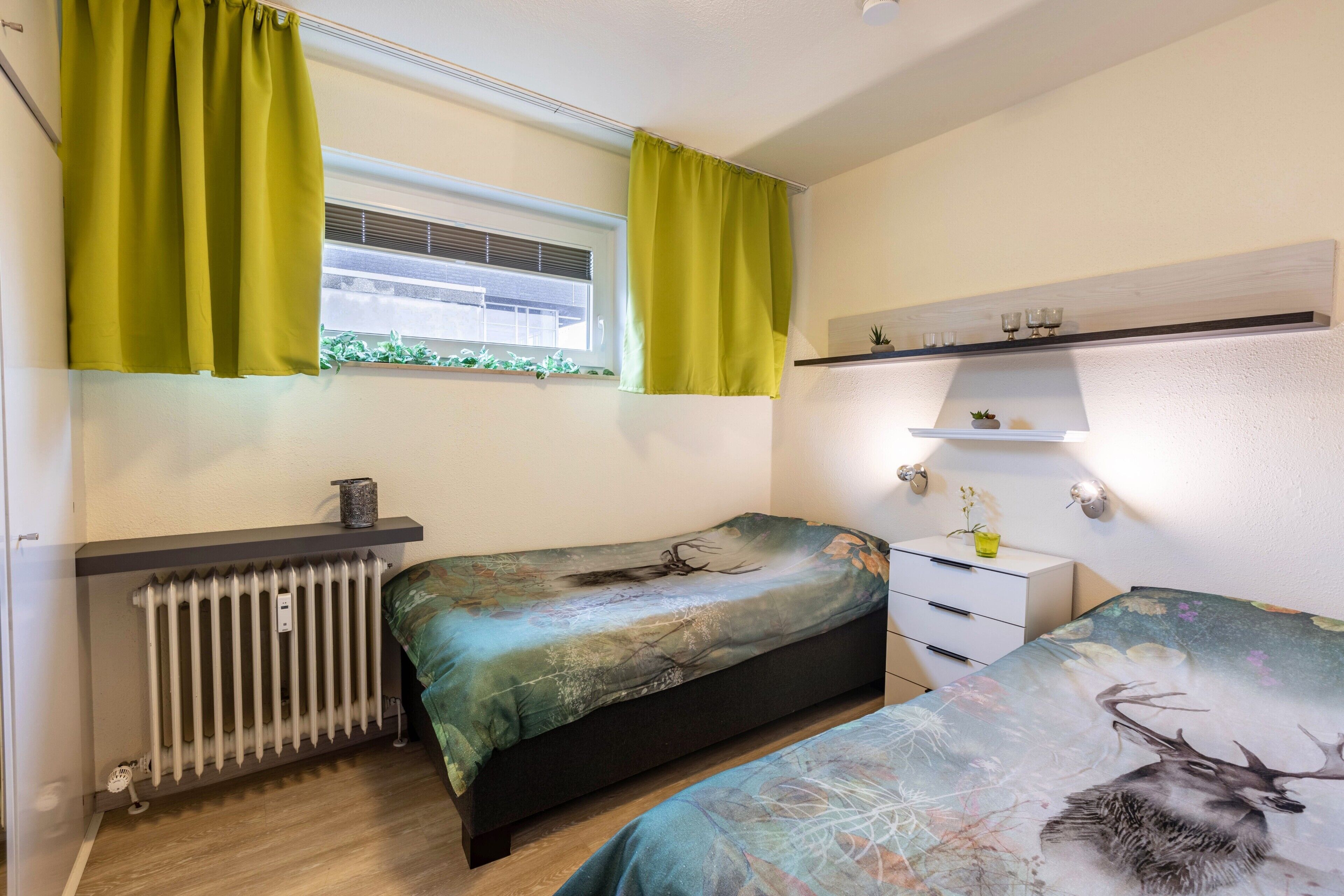 Een slaapkamer met twee bedden, een radiator, een raam met gordijnen, een plank met decoratieve objecten en een kleine plant.