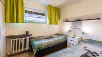 Een slaapkamer met twee bedden, een radiator, een raam met gordijnen, een plank met decoratieve objecten en een kleine plant.