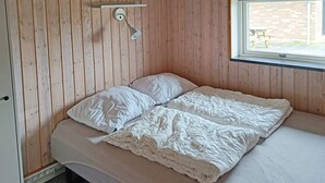 Un lit simple avec une couette et des oreillers blancs, un mur en bois, une fenêtre donnant sur l’extérieur et un luminaire fixé au mur.