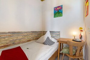 Ein Einzelbett mit roter Decke, ein hölzerner Kopfteil, ein kleiner Tisch mit Lampe und ein Gemälde an der Wand.
