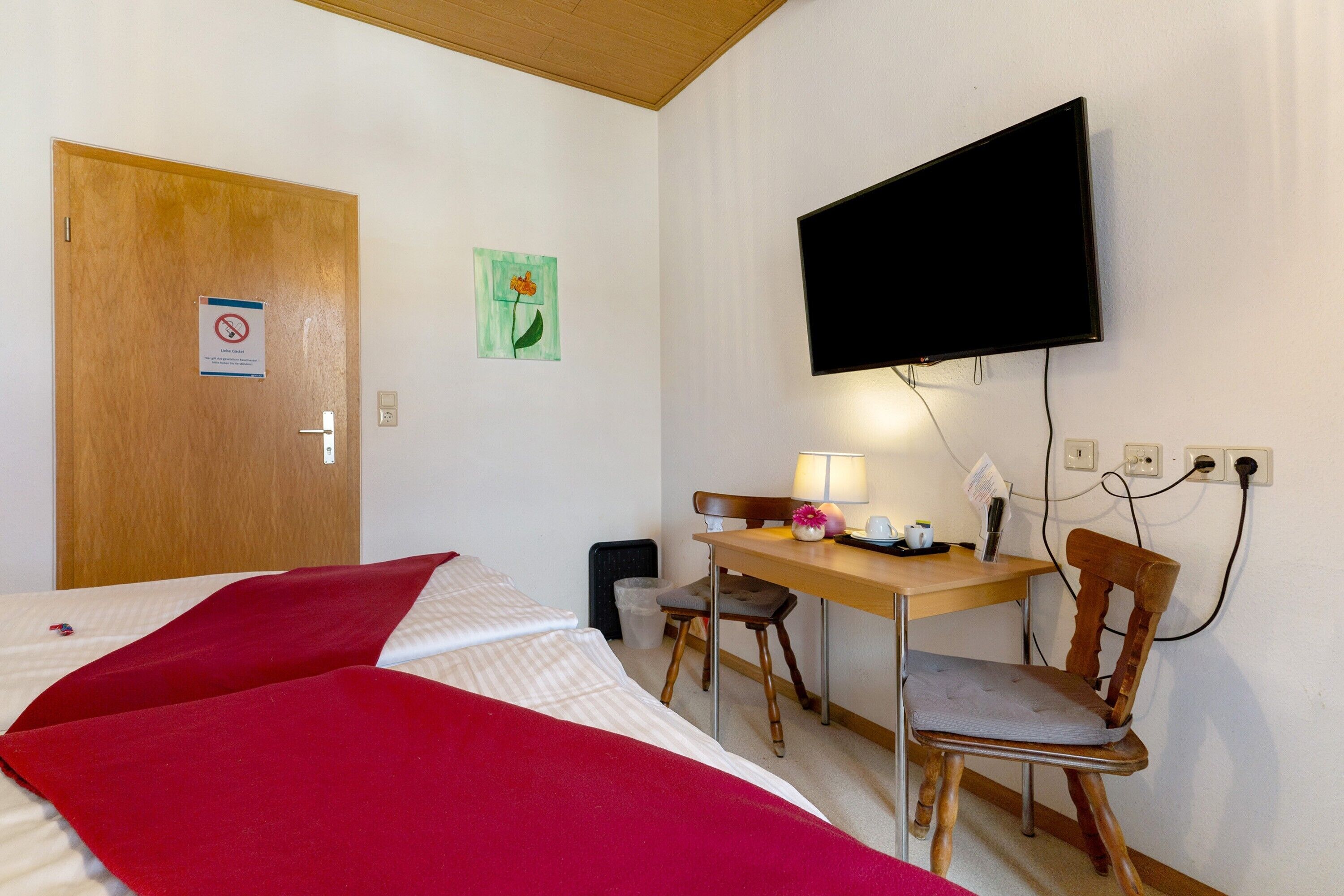 A hotel room with a bed, a desk with a chair, a television mounted on the wall, and a door with a sign.