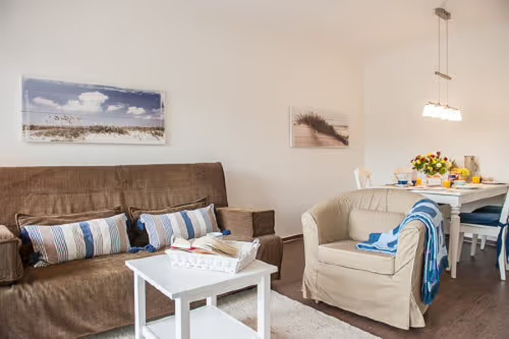 Una sala de estar con un sofá marrón, una mesa de centro blanca, un sillón beige y un comedor con una mesa y sillas.