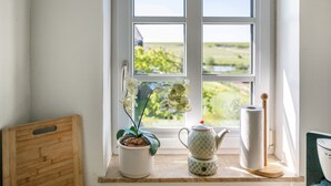 Et vindue med udsigt over et grønt landskab, en potteplante, en tepotte og en træskærebræt på et vindueskarm.